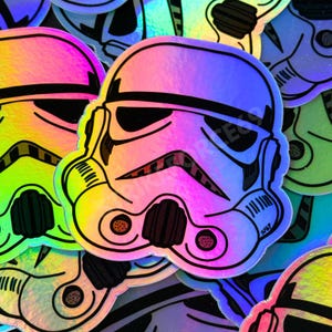 May include: A holographic sticker featuring a stylised black and white illustration of a stormtrooper helmet. The design includes bold black outlines and a reflective, rainbow-coloured surface that shifts in light, showcasing the iconic facial features.