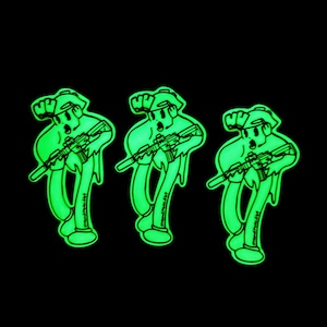 May include: Three green glow-in-the-dark stickers featuring a cartoon character holding a rifle. The character is wearing a hat and has a surprised expression. The text "madvillain" is printed on each sticker.