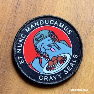 May include: A black and red embroidered patch with a cartoon seal wearing a helmet and eating spaghetti. The text on the patch reads "Et nunc manducamus gravy seals".