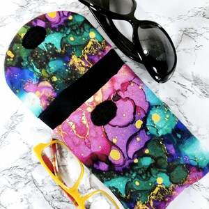 May include: A colorful glasses case with a vibrant abstract design in shades of purple, teal, and gold. The case is open, revealing a black interior. Black sunglasses and yellow-framed glasses are also visible.