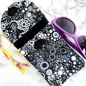 May include: A black and white patterned glasses case with a magnetic closure, next to yellow and purple sunglasses. The case features a repeating circle design in various sizes.