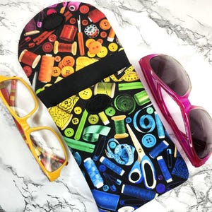 May include: A black eyeglass case with a colorful sewing-themed pattern, featuring spools of thread, buttons, and scissors. A pair of yellow eyeglasses and pink sunglasses are also in the image.