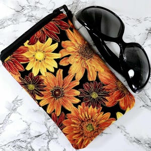 May include: A black zippered pouch with a floral print featuring orange, yellow, and red sunflowers on a black background. The pouch is lying on a white marble surface next to a pair of black sunglasses.