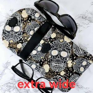 May include: Black and white fabric eyeglasses case with a sugar skull design. The case has a black strap and a black lining. The case is extra wide.
