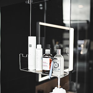 May include: A clear plastic shower caddy with four bottles of toiletries. The caddy is hanging from a shower head. The bottles are white, clear, and brown. The bottles have labels with text. A white bath sponge is hanging below the caddy.