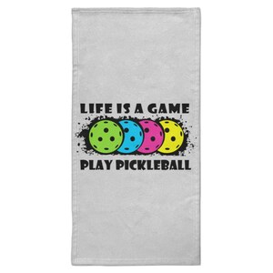 Può includere: Un asciugamano grigio chiaro con la scritta "LIFE IS A GAME PLAY PICKLEBALL". L'asciugamano presenta quattro palline da pickleball verdi, blu, rosa e gialle. Le palline da pickleball sono sopra il testo "PLAY PICKLEBALL".
