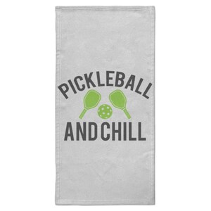Può includere: Un asciugamano grigio chiaro con le parole "PICKLEBALL AND CHILL" in nero, con racchette da pickleball e una palla verdi. L'asciugamano è rettangolare e sembra essere fatto di un materiale morbido.
