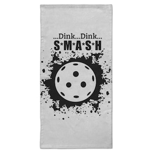 Può includere: Un asciugamano grigio chiaro con una grafica nera. La grafica presenta un disegno a palla da pickleball con le parole "...Dink...Dink...S*M*A*S*H" in grassetto nero. La palla è circondata da un disegno a spruzzi neri.