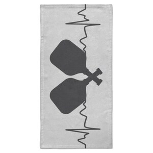 Può includere: Un asciugamano grigio chiaro con un design grafico nero. Il design include due racchette da pickleball incrociate, con una linea di battito cardiaco verticale. L'asciugamano è rettangolare.