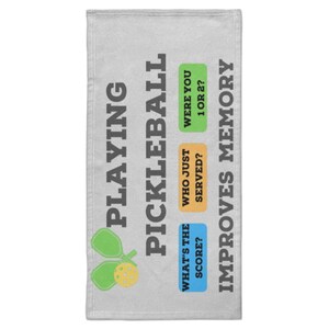 Può includere: Un asciugamano grigio chiaro con le parole "PLAYING PICKLEBALL IMPROVES MEMORY" in grigio scuro. L'asciugamano presenta una grafica di pickleball e tre riquadri colorati con le domande: "WHAT'S THE SCORE?", "WHO JUST SERVED?" e "WERE YOU 1 OR 2?"