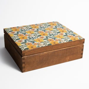 Può includere: Una scatola di legno con un motivo floreale sul coperchio. Il coperchio presenta un motivo di fiori gialli, foglie verdi e accenti bianchi su uno sfondo chiaro. La scatola è in legno marrone.