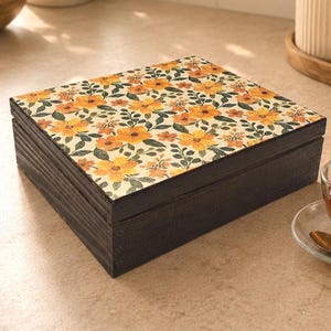 May include: A dark brown wooden box with a floral design on the lid. The lid features a pattern of yellow and orange flowers with green leaves on a cream background. A cup of tea, saucer, and spoon are in the foreground.