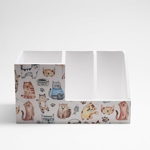 Peut inclure: Organiseur de bureau en bois blanc avec quatre compartiments. L'organisateur est décoré d'un motif de chats et d'empreintes de pattes. Le mot "chat" est imprimé sur le devant de l'organisateur.