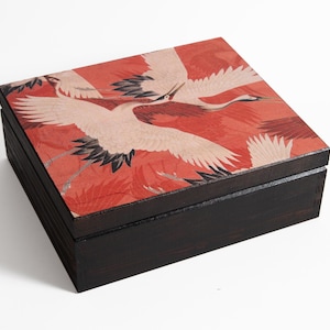 May include: A square wooden box. The lid has a red background with white and red cranes in flight. The box has a dark brown base. Likely used for storage or decoration.