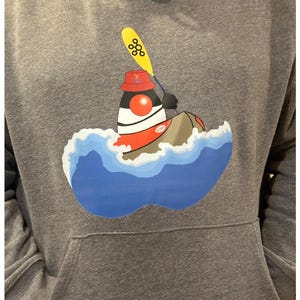 May include: Grey hooded sweatshirt with a front pocket. The sweatshirt features a colourful graphic of a cartoon character in a kayak, paddling through blue and white waves. The character wears a red hat and holds a yellow paddle.