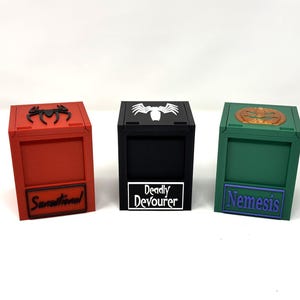 Puede incluir: Tres cajas cuadradas de colores con tapas con bisagras. La caja roja tiene un emblema de araña negro y la palabra "Sensational" en la parte delantera. La caja negra tiene un emblema de araña blanco y las palabras "Deadly Devourer". La caja verde tiene un emblema circular y la palabra "Nemesis".