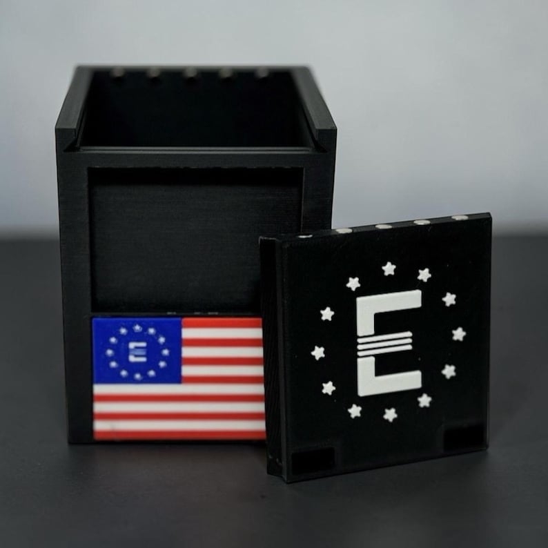 Pu&ograve; includere: Una scatola di legno nera con coperchio rimovibile. La scatola presenta un piccolo disegno della bandiera americana e un coperchio con un logo "E" bianco circondato da stelle. La scatola &egrave; esposta su una superficie scura.