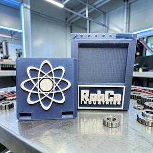 Pu&ograve; includere: Due scatole blu stampate in 3D. Una presenta un simbolo di atomo bianco, l'altra un'etichetta con la scritta "RobCo INDUSTRIES" in bianco. Le scatole sono su una superficie metallica con ingranaggi.