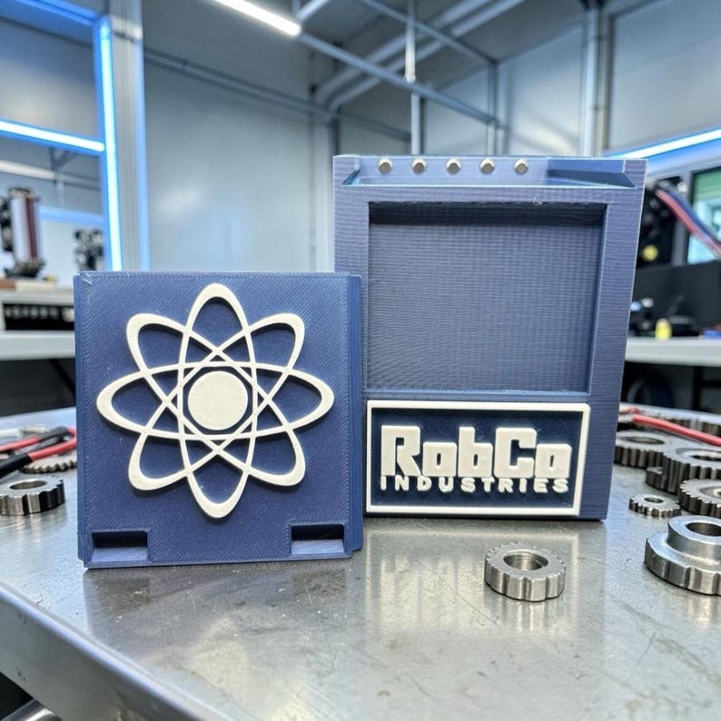Pu&ograve; includere: Due scatole blu stampate in 3D. Una presenta un simbolo di atomo bianco, l'altra un'etichetta con la scritta "RobCo INDUSTRIES" in bianco. Le scatole sono su una superficie metallica con ingranaggi.