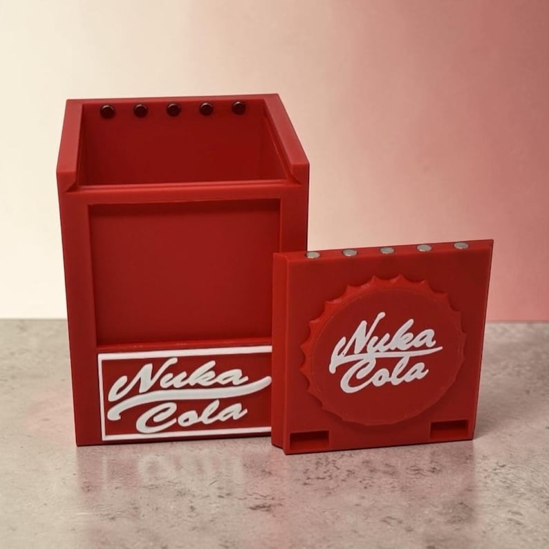 Pu&ograve; includere: Un organizer da scrivania rosso e un piccolo contenitore quadrato, entrambi con il logo "Nuka Cola" in bianco. L'organizer da scrivania ha una forma rettangolare con un ritaglio e il contenitore quadrato assomiglia a un tappo di bottiglia.
