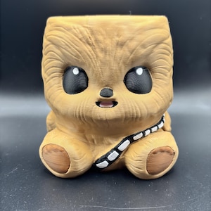 May include: A brown and white Chewbacca character planter with a black bandolier. The planter has a square base and round feet. The character has large eyes and a small mouth.