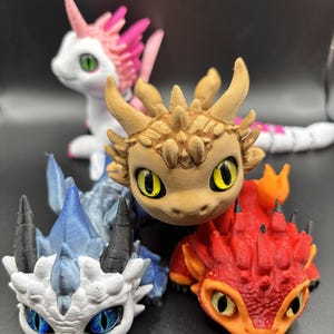 Puede incluir: Cuatro pequeñas figuritas de dragón impresas en 3D, de colores vivos. Los dragones están en varias poses, uno de pie sobre sus patas traseras, otro acostado y dos sentados. Los dragones son todos de diferentes colores, uno es rosa, otro marrón, otro azul y otro rojo.