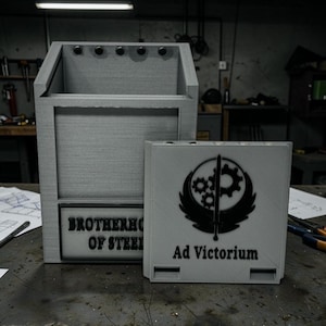 Pu&ograve; includere: Una scatola portaoggetti grigia con la scritta "BROTHERHOOD OF STEEL" e una scatola grigia pi&ugrave; piccola con la scritta "Ad Victorium" e un emblema nero. Le scatole sono su una superficie metallica.
