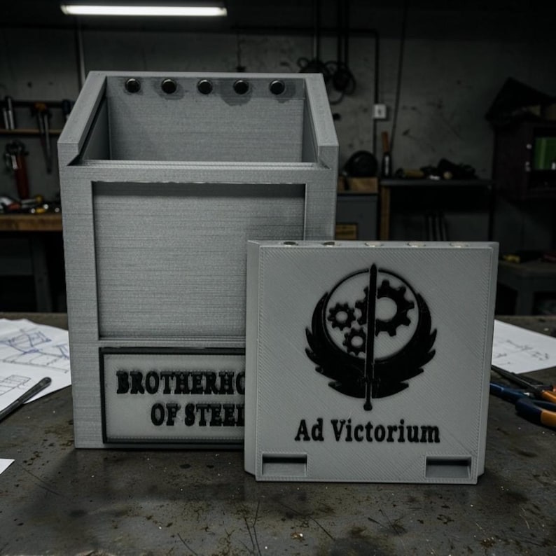 Pu&ograve; includere: Una scatola portaoggetti grigia con la scritta "BROTHERHOOD OF STEEL" e una scatola grigia pi&ugrave; piccola con la scritta "Ad Victorium" e un emblema nero. Le scatole sono su una superficie metallica.