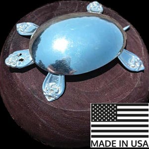May include: A silver turtle sculpture made from repurposed silverware. The turtle is sitting on a dark brown wooden surface. The sculpture has a shiny, reflective finish. Made in the USA.