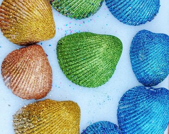 Op de afbeelding: Een bovenaanzicht van verschillende met glitter bedekte schelpen in goud, koper, groen en blauw. De schelpen zijn gerangschikt op een witte ondergrond, met een paar glitterspikkels verspreid. De schelpen hebben een getextureerd, geribbeld uiterlijk.
