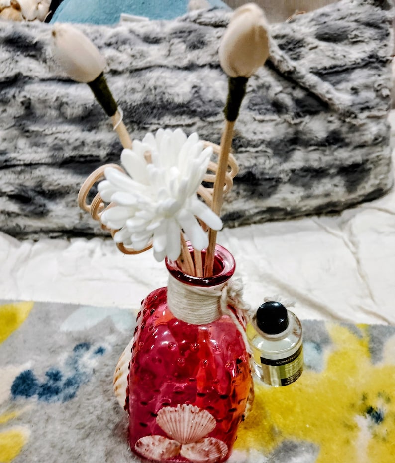 Op de afbeelding: Een rode glazen diffuserfles met een witte bloem en houten stokjes. De fles heeft een getextureerd oppervlak en is versierd met schelpen. De diffuser staat op een kleurrijk tapijt.