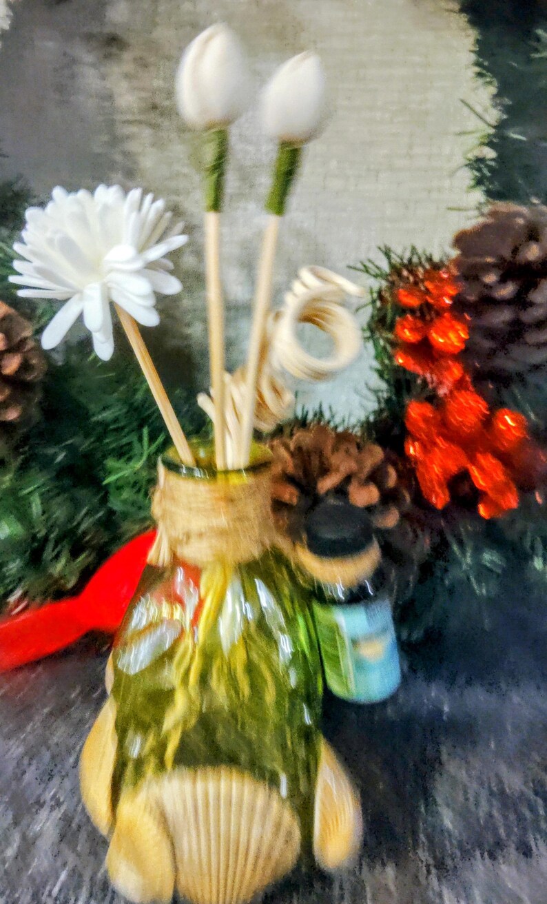 Op de afbeelding: Een heldere glazen diffuserfles met een jutewikkel en een schelpbasis. De fles is gevuld met houten stokjes en witte bloemenaccenten.