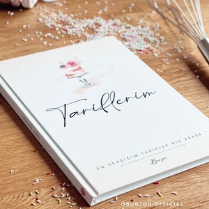 Könnte beinhalten: Ein weißes Kochbuch mit dem Titel "Tariflerim" in schwarzer Kursivschrift. Das Cover zeigt eine Aquarell-Illustration eines rosa und weißen Kuchens mit Blumen darauf. Der Text unter dem Titel lautet "En Sevdigim Tarifler Bir Arada" und "Burjou". Am unteren Rand des Covers steht der Text "@burjou_official".