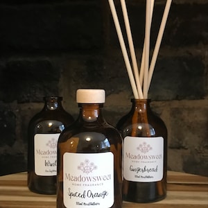 May include: Three brown glass bottles with labels and white caps. The bottles contain reed diffusers with scents of Spiced Orange, Gingerbread, and Want. The bottles are sitting on a wooden surface.