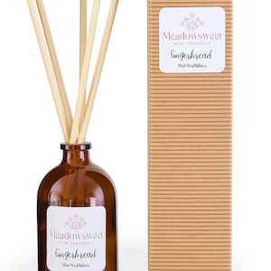 May include: A brown glass bottle with a white label that reads "Meadowsweet Home Fragrance Gingerbread 100ml Reed Diffuser". The bottle is filled with a clear liquid and has a set of brown wooden sticks inserted into the top. The bottle is sitting on a white surface next to a brown cardboard box with the same label.