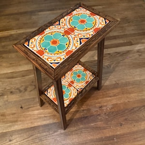 May include: A rectangular wooden side table featuring two tiers, each inlaid with decorative ceramic tiles. The tiles display a repeating floral pattern in vibrant shades of turquoise, orange, and yellow, set within a dark brown wooden frame.