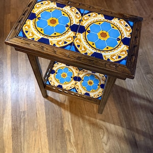 May include: A rectangular wooden side table featuring a dark brown frame with carved edges. The top and lower shelf are each inlaid with two square ceramic tiles displaying a vibrant blue, yellow, and white geometric floral pattern.