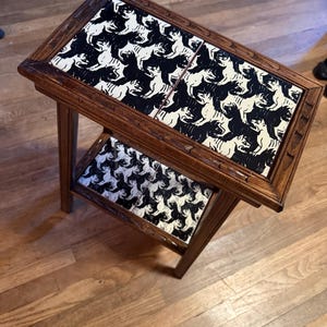 May include: A rectangular wooden side table with four legs and two tiers. The top and bottom surfaces feature inlaid square tiles decorated with a repeating black and white graphic pattern of interlocking dog silhouettes in a tessellated design.