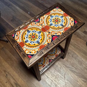 May include: A rectangular wooden side table with a decorative tile top. The tiles feature a repeating pattern of yellow, blue, orange, and red floral designs. The table has a lower shelf with matching tiles and a dark brown finish.