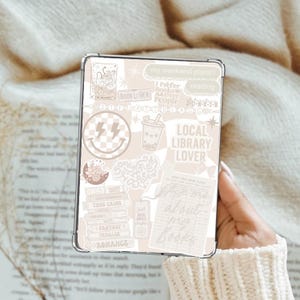 May include: A clear phone case with a beige and white sticker collage. The stickers include text like "Book Lover", "Local Library Lover", and "my weekend plans?" along with various graphics such as a smiley face and a boba drink.