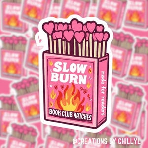May include: Pink and white matchbox sticker with the text "Slow Burn" and "Book Club Matches" in black. The matchbox has a red and yellow flame illustration and pink hearts. The text "made for readers" is on the right side of the matchbox.