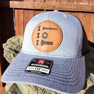 May include: A gray and black trucker hat with a brown leather patch featuring a screw, nut, and bolt. The patch has the text "I I I" above the hardware. The hat has a black mesh back and a black adjustable strap with a black label that reads "EST. 1970 RICHARDSON. ORIGINAL 112 TRUCKER ADJUSTABLE."