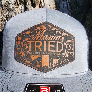 May include: A gray and blue trucker hat with a black and brown leather patch that says "Mama Tried" and "Only Hell Mama Raised".