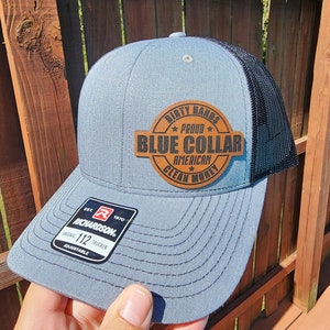 May include: A gray and black trucker hat with a brown leather patch that says "Dirty Hands, Proud, Blue Collar, American, Clean Money". The hat is adjustable and has a Richardson logo.