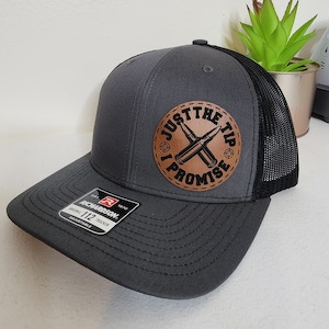 May include: A gray and black trucker hat with a brown leather patch that says "Just the Tip I Promise" with a crossed bullet design.