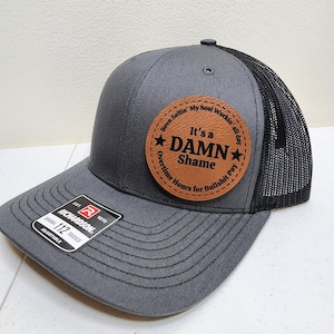 May include: Gray trucker hat with a black mesh back and a brown leather patch. The patch features the text "It's a DAMN Shame" and other phrases. The hat is adjustable and has the Richardson logo.