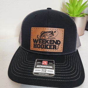 May include: Black and gray trucker hat with a brown leather patch that says "Weekend Hooker" with a fish graphic. The hat is adjustable and has a Richardson 112 label.
