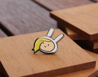 Puede incluir: Un colorido pin de esmalte con un personaje de dibujos animados con una capucha blanca con orejas de conejo y pelo rubio. El pin está colocado sobre un cuadrado de madera, con otros cuadrados de madera en el fondo.