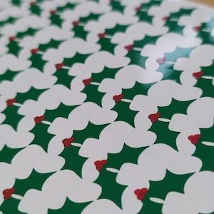May include: A repeating pattern on a smooth surface featuring white holly leaf shapes layered over a dark green background, accented with small clusters of three red berries arranged in a consistent grid across the entire visible area.