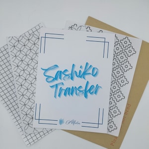 Puede incluir: Un conjunto de cuatro patrones de transferencia Sashiko en papel blanco. Los patrones son de diseño geométrico en blanco y negro. El papel está apilado sobre un sobre marrón. El texto "Sashiko Transfer" está escrito en azul en la hoja de papel superior.